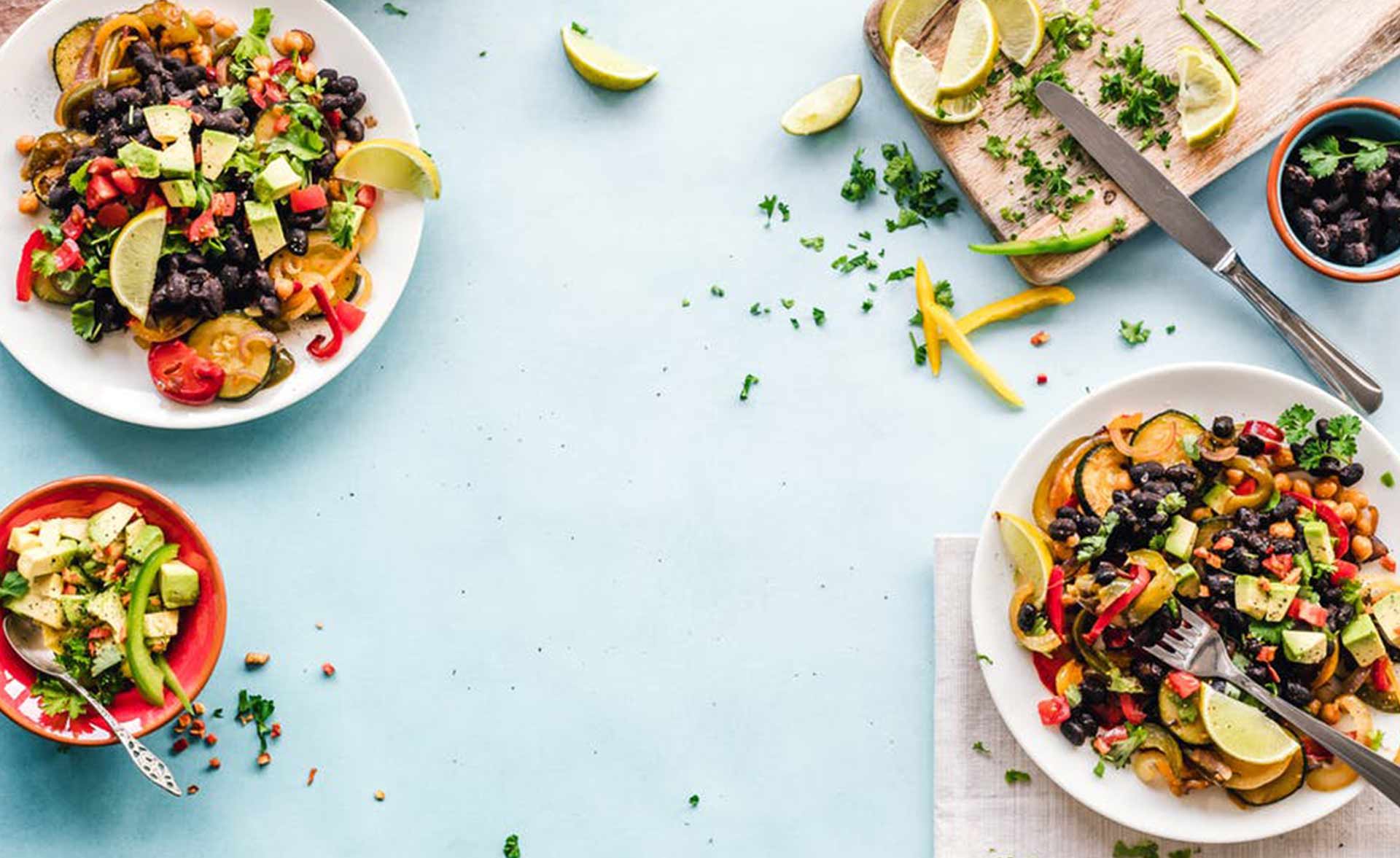 Three plates of colorful salad with a variety of vegetables, topped with lime wedges and beans, placed on a light blue table. A cutting board with a knife, some chopped vegetables, and a small bowl of beans are seen in the background. Lime wedges and herbs are scattered around.