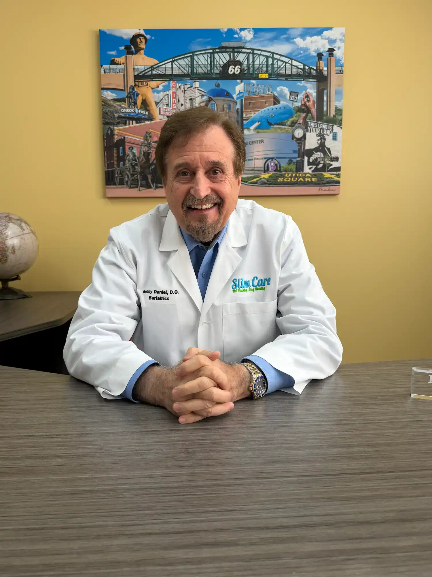 Dr Daniels, owner and CEO of Slim Care Medical Weight Loss Clinic, sits at a desk smiling in a white coat with the Slim Care logo, with a colorful Tulsa mural behind him highlighting the clinic’s local roots.