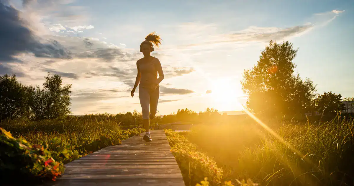 Woman jogging along a wooden path at sunset with headphones on as golden sunlight shines through tall grass and trees. This outdoor workout scene reflects healthy habits and motivation tied to summer weight loss in Tulsa.