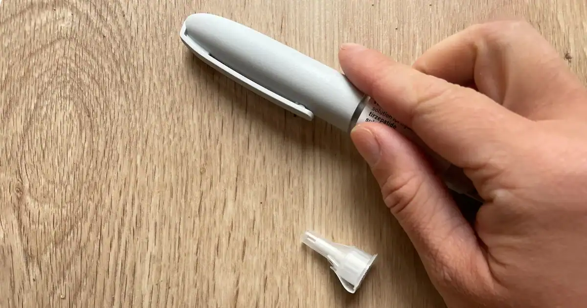 A hand holding a gray injection pen with a white cap labeled "tirzepatide" on a wooden surface, next to a detached plastic needle cap.